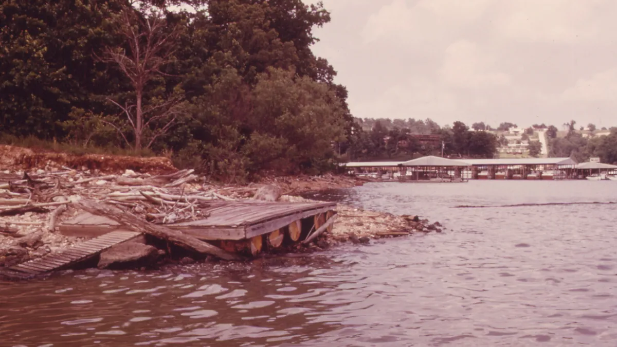 An old wooden dock washed up on a rocky shoreline, with covered marina docks across the water.
